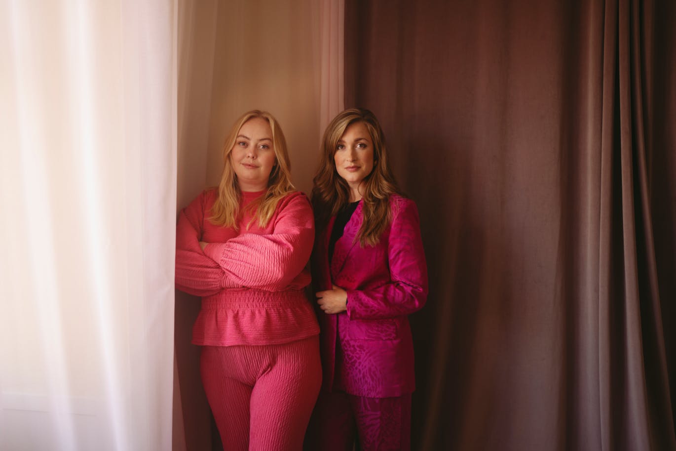 Maria Gudme og Camilla Søe står sammen i et rum med gardiner i lyse farver. Maria Gudme står forest i et rødt sæt tøj, Camilla Søe står bag hende i et pink jakkesæt.