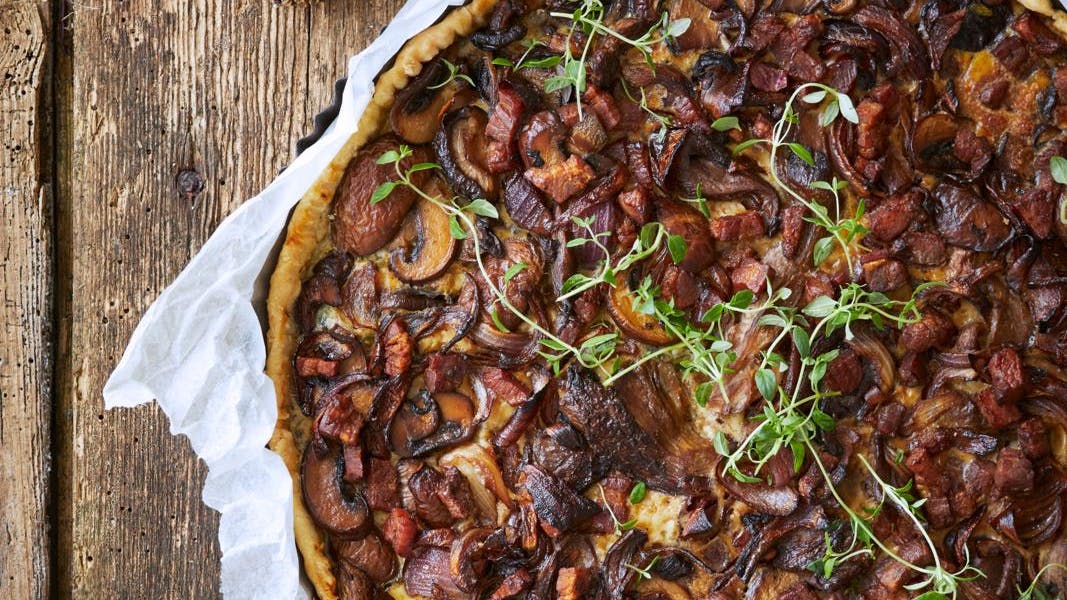 Svampetærte med karamelliserede løg og bacon serveret på rustik bordplade.