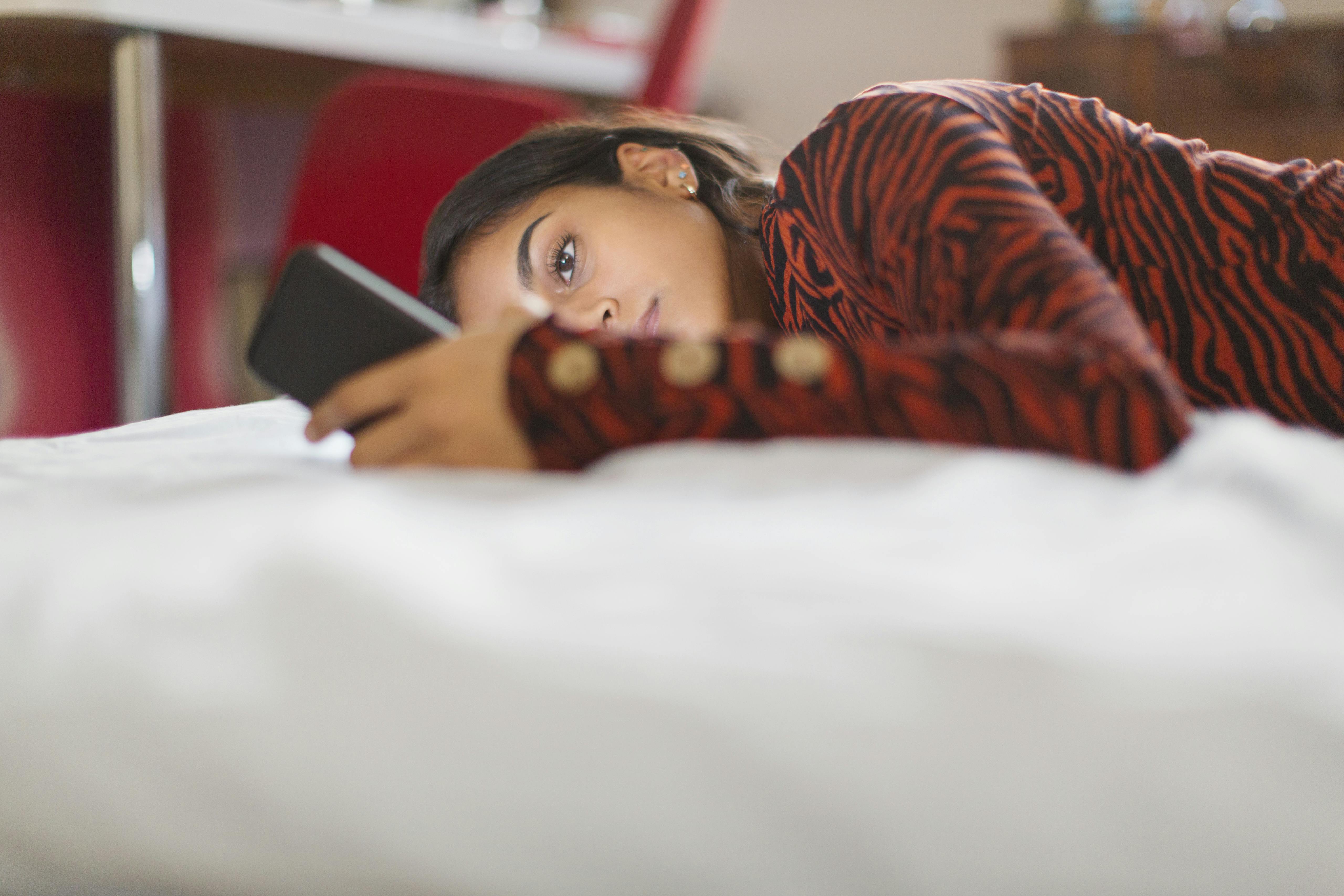 Focused teenage girl using smart phone laying on bed.