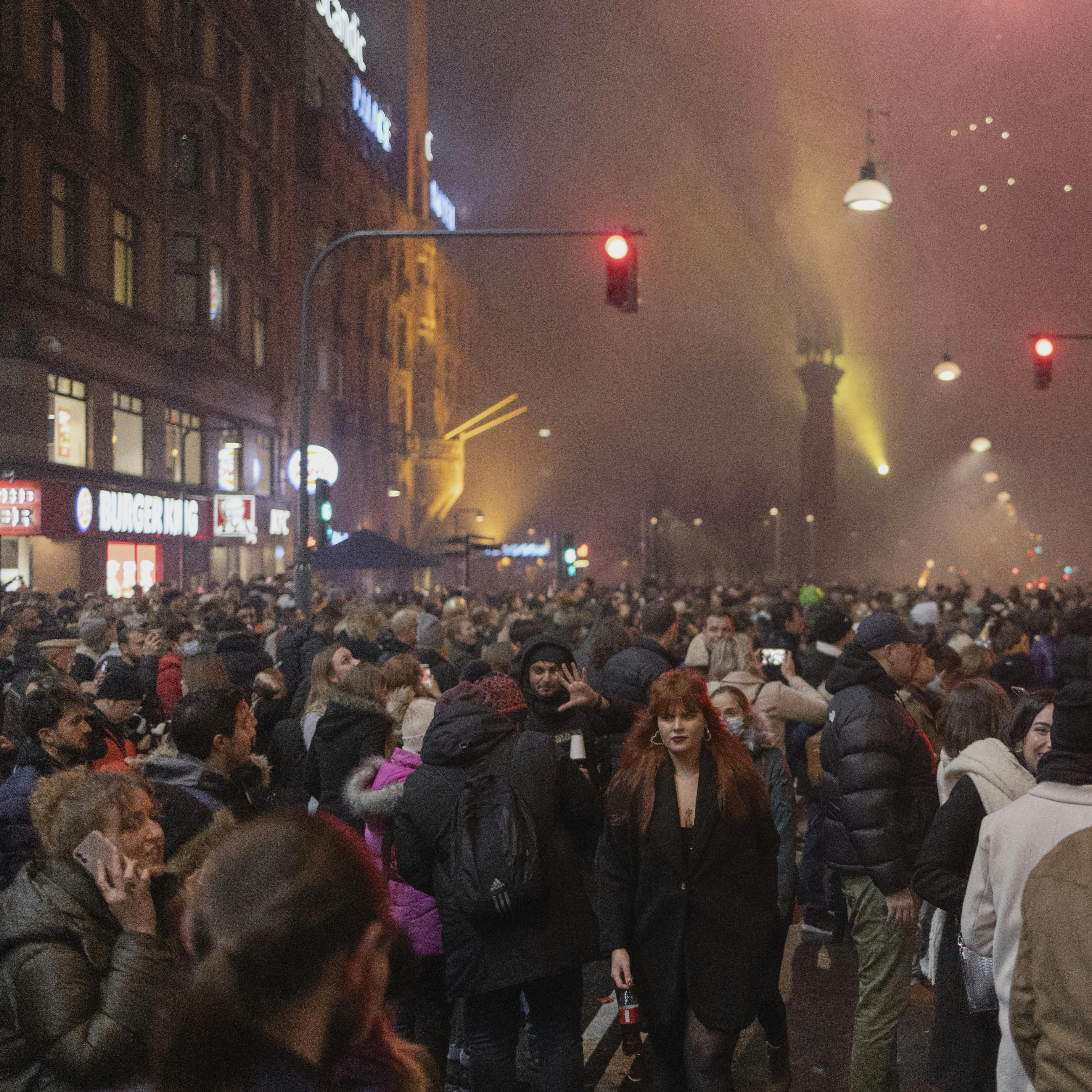 Nytårsaften 2021 - 2022. I et kollegiekøkken blev der skålet højt i farvede shotsglas. En corona-ramt familie så dronningens nytårstale under sølvfarvede serpentiner. To veninder måtte udskyde nytårs-byturen et år mere, men på Rådhuspladsen fløj raketterne om ørerne, som de plejer. Mennesker og fyrværkeri på Rådhuspladsen.