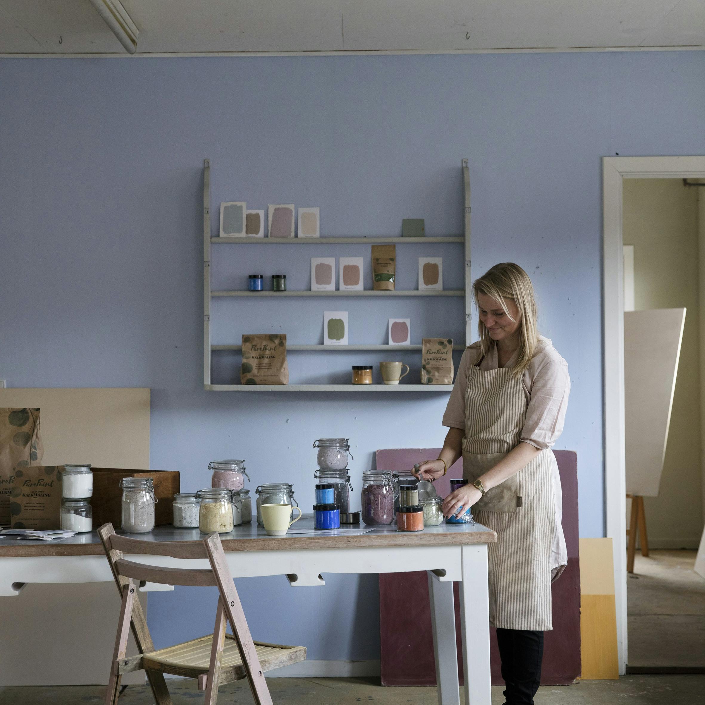 Johanne med forklæde ved det lange arbejdsbord i sit værksted, der er malet i Forglemmigej Blå.