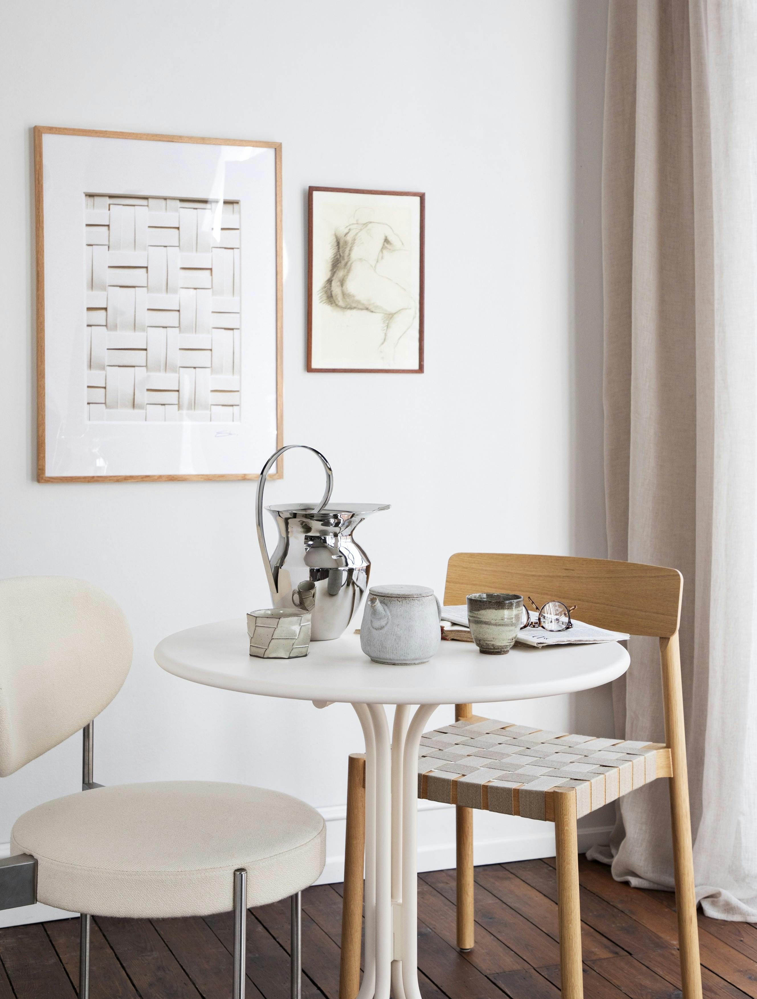 Cafebord og stol med fletsæde fra &Tradition, polstret stol fra Verpan, kande fra Audo Copenhagen, krus fra Sakura Copenhagen, gardin fra Jysk og plakat i ramme fra Desenio.