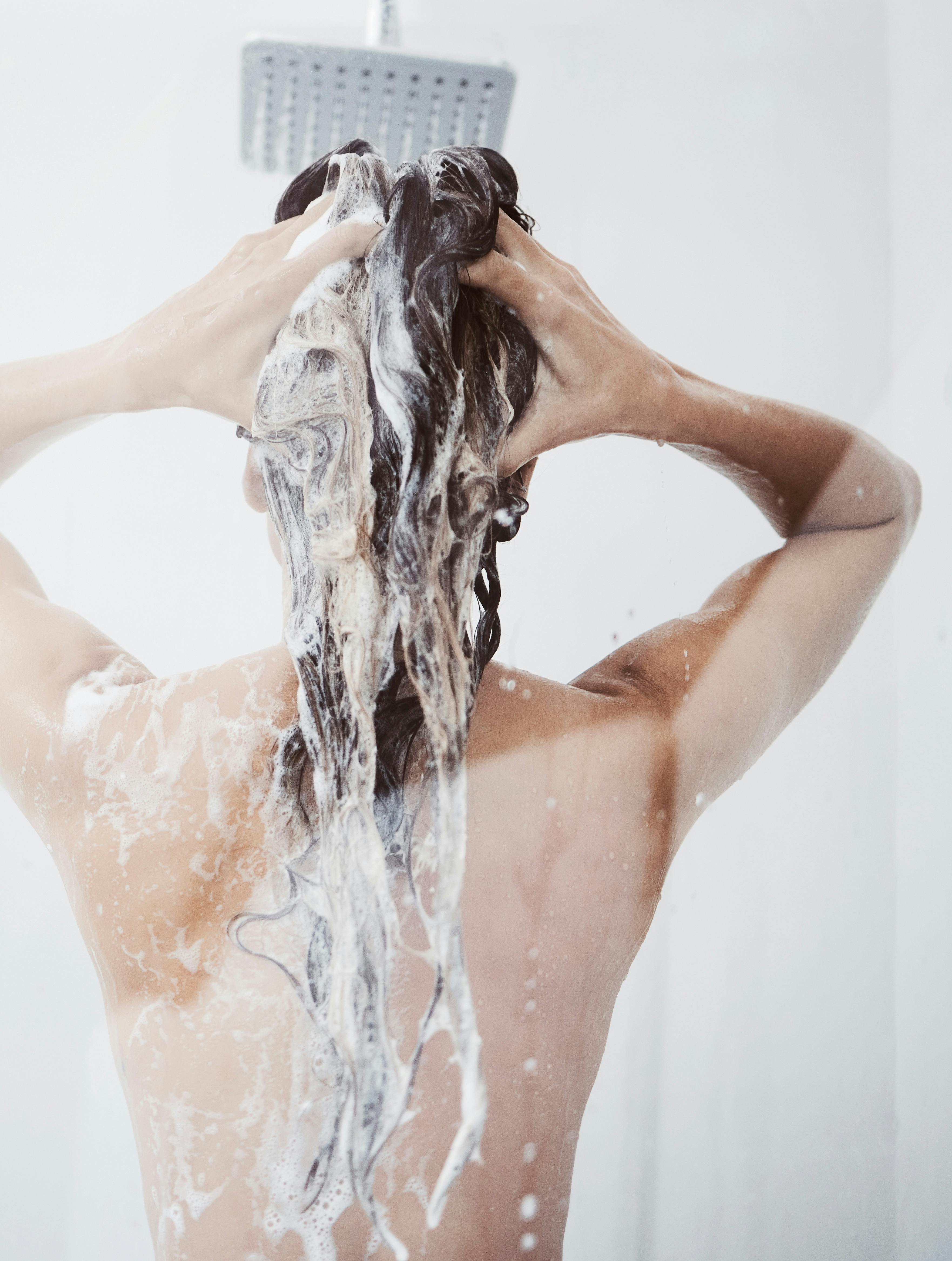 Shower, washing and woman in bathroom with haircare for shampoo, natural treatment or organic cosmetics. Back view, body and female person with foam for hygiene, cleaning or morning routine in home.