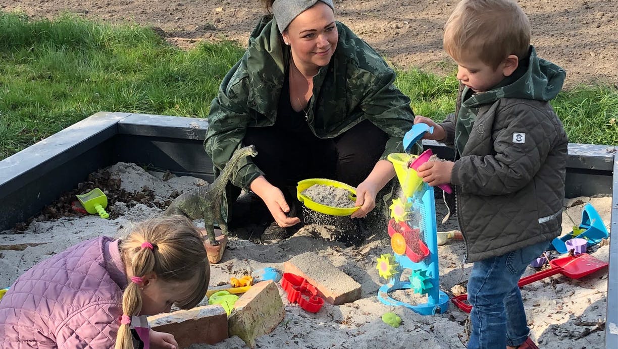 Familie leger i sandkasse mens mor hjemmepasser